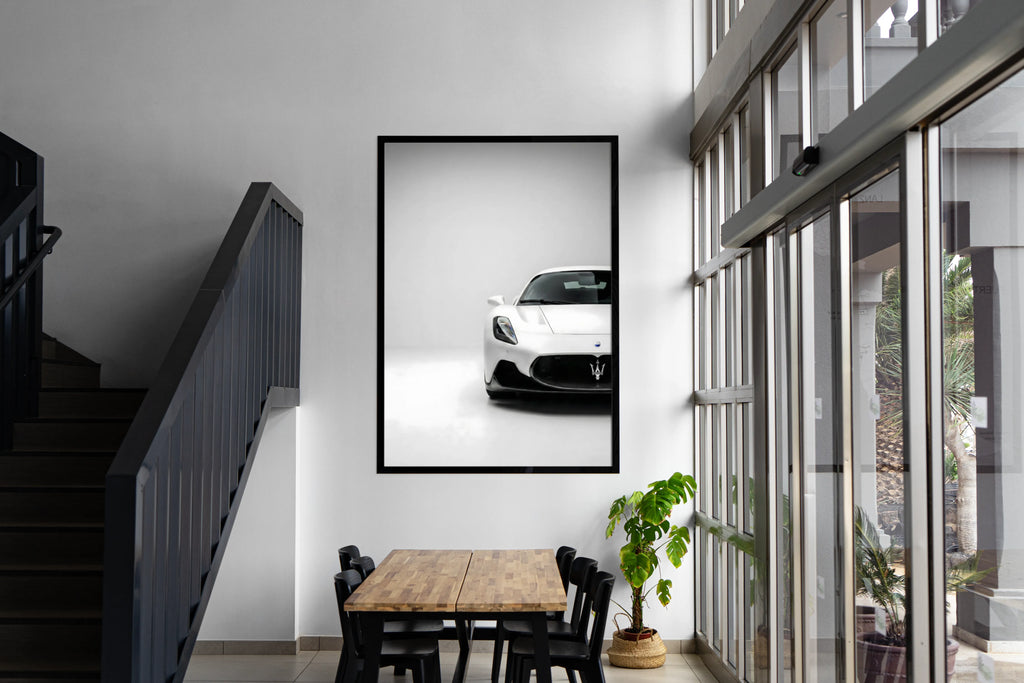 Framed picture of a white car on a wall above a dining table with a staircase and large window in the background.