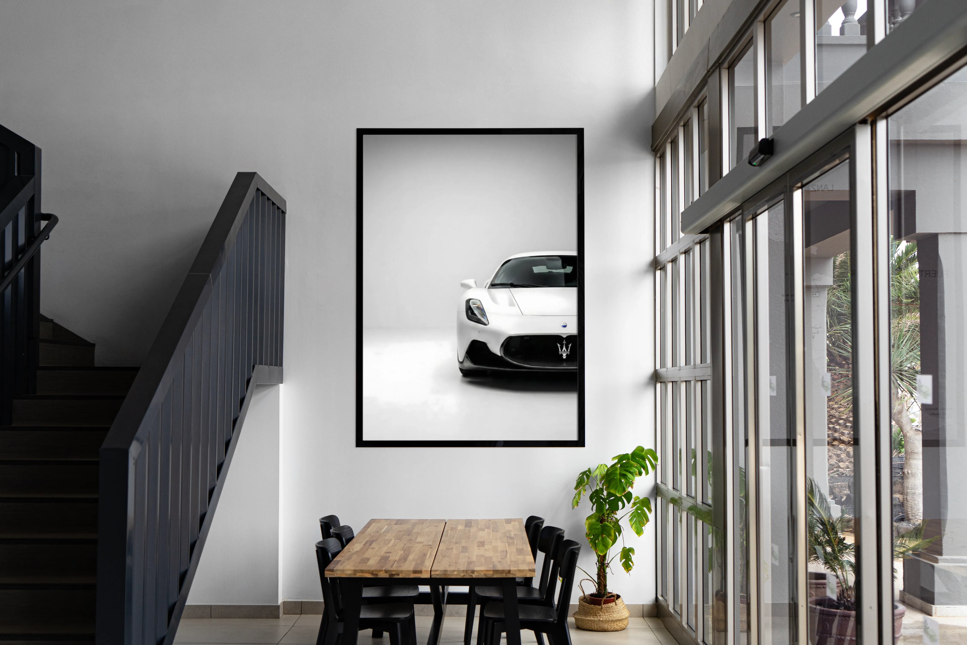 Framed picture of a white car on a wall above a dining table with a staircase and large window in the background.