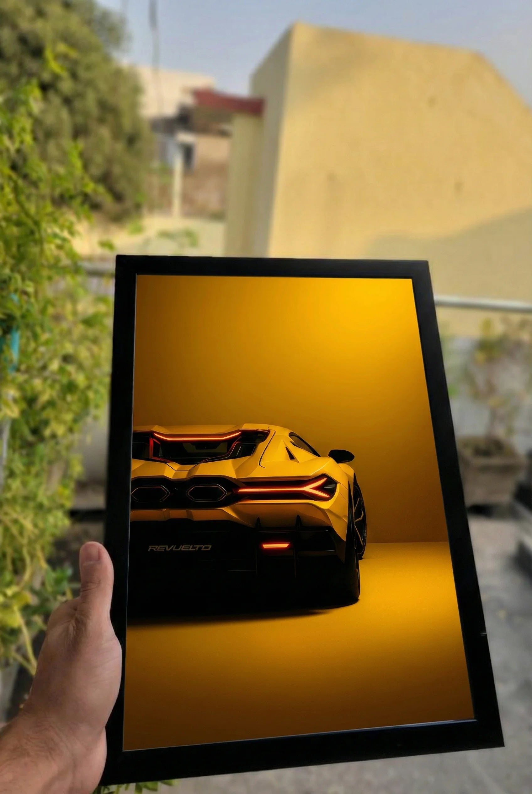 Framed picture of a yellow sports car with red interior held by a hand outdoors.